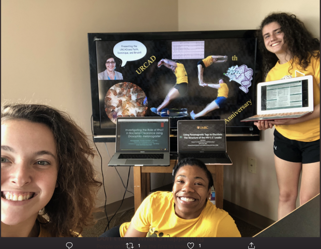Three young women smile at the camera. One is close to the camera, another sits on the floor behind her, and the third is in the background standing holding a laptop. There is a large monitor and two lap tops with images behind them.