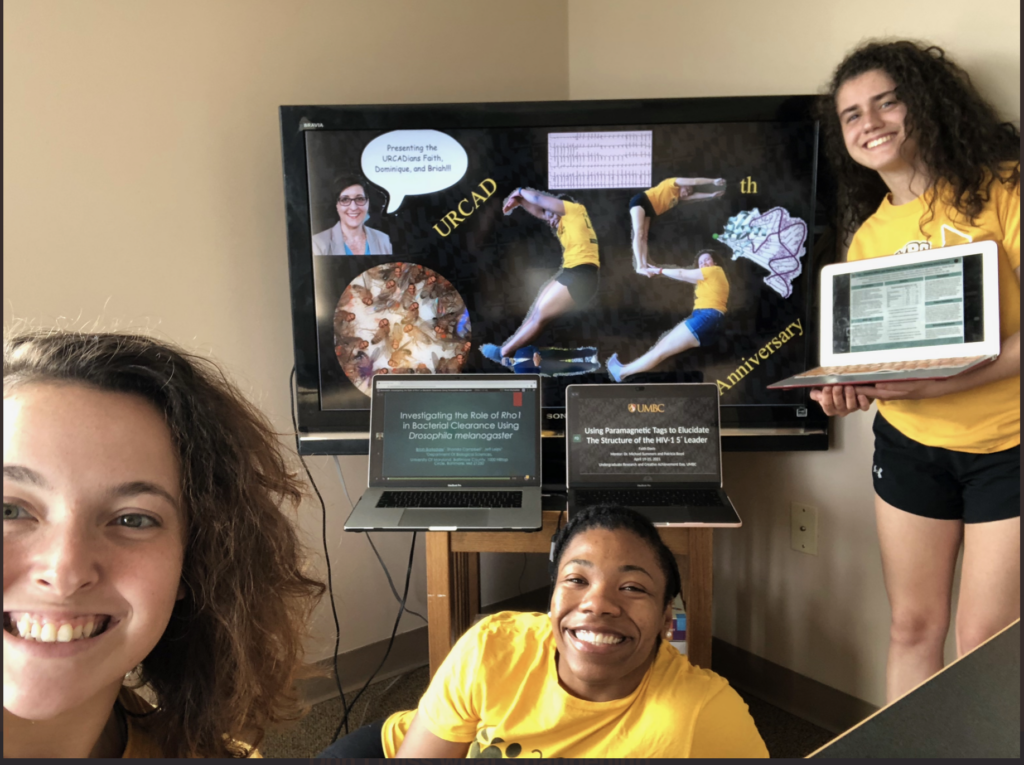 Three young women smile at the camera. One is close to the camera, another sits on the floor behind her, and the third is in the background standing holding a laptop. There is a large monitor and two lap tops with images behind them.