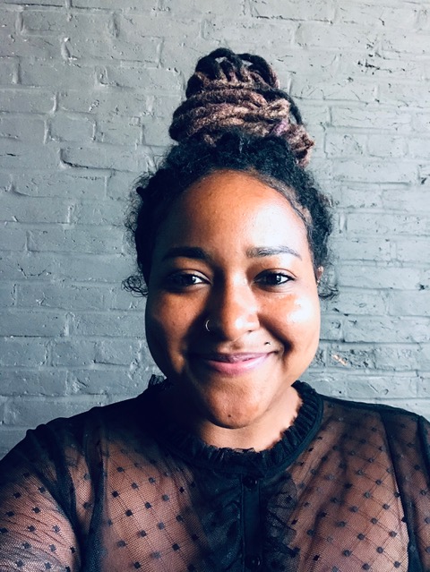 A young woman with black hair pulled back high on top of her head with a scrunchy and wearing a black blouse with dots in a diamond pattern smiles at the camera.