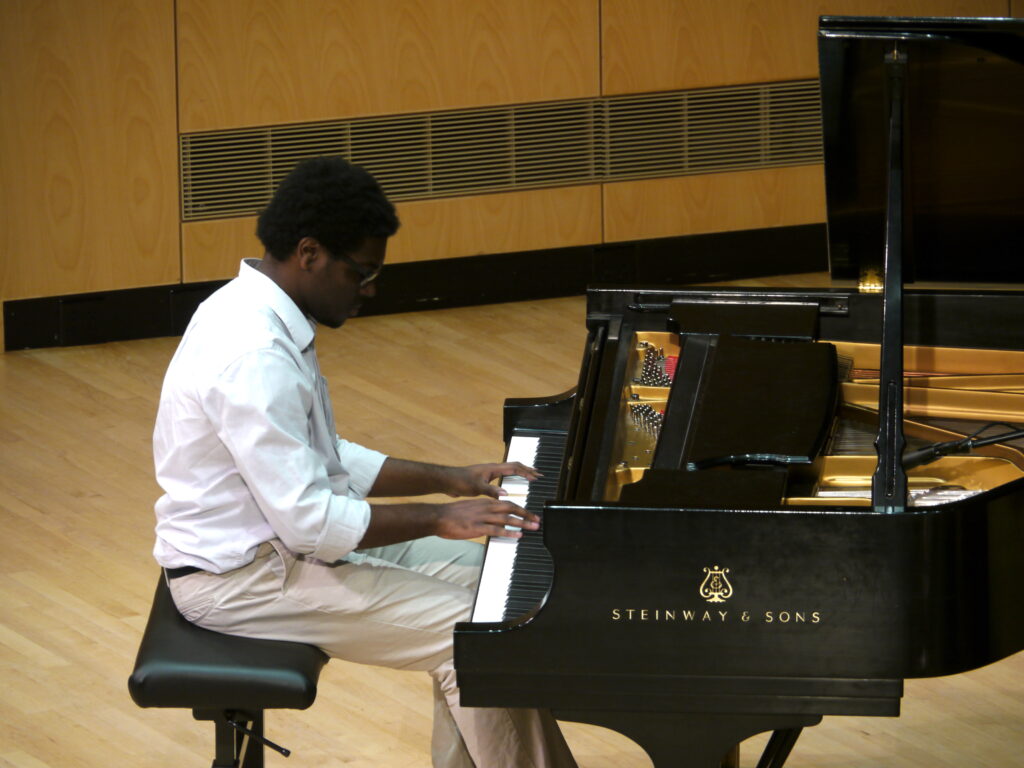 A young man plays a grand piano in a concert hall.