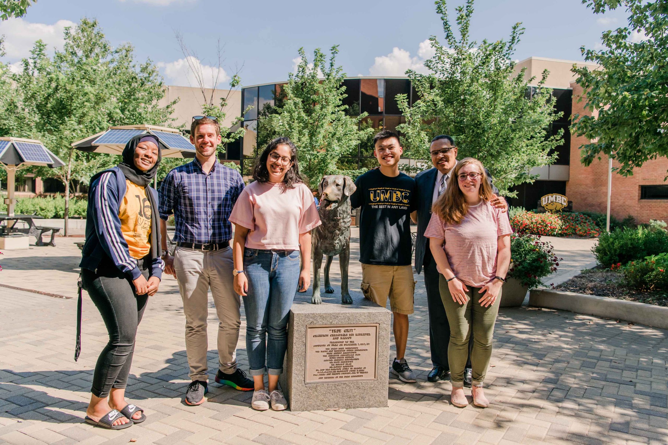 Six people stand in the sunshine, posing with a statue of a dog.