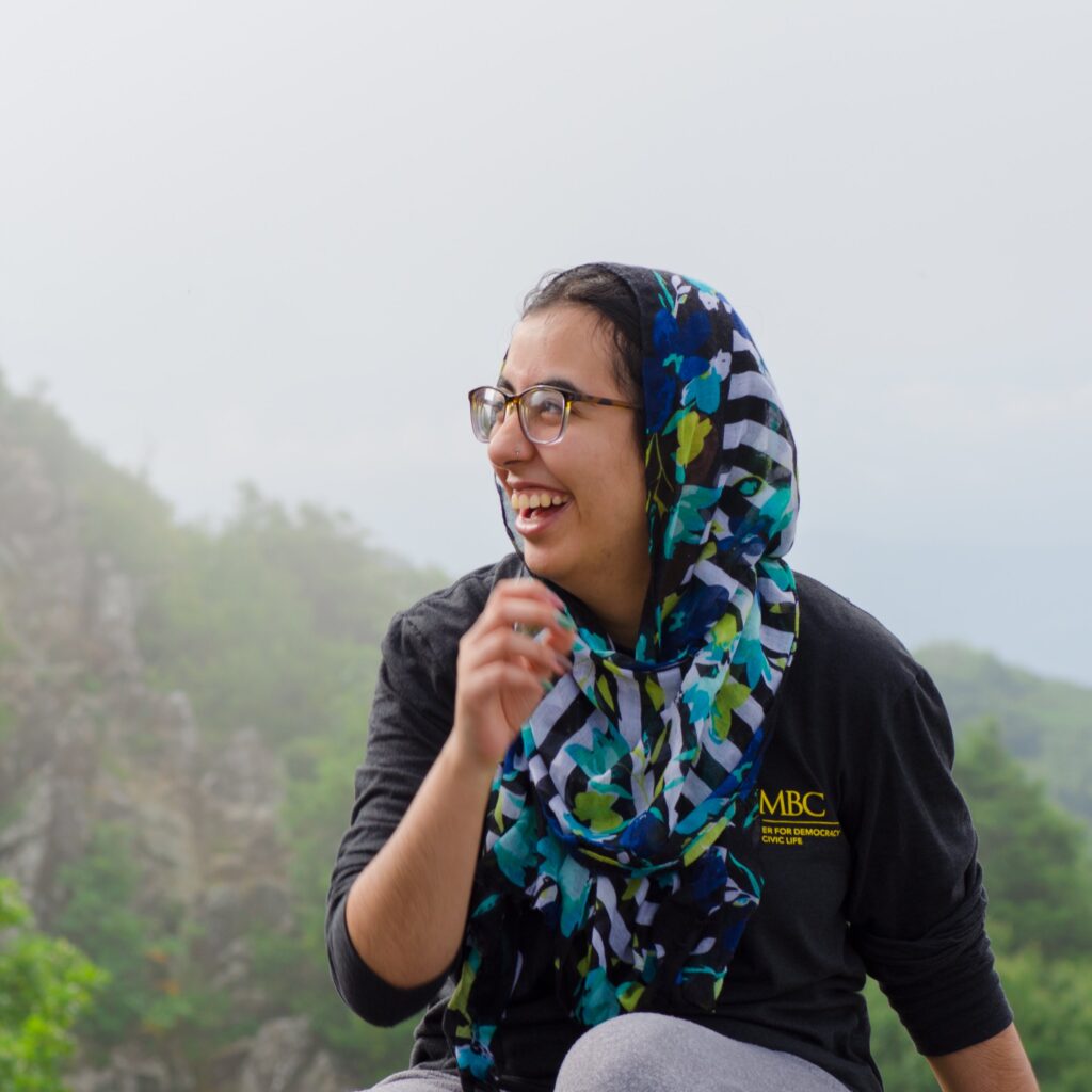 Young women sits outside, smiling, wearing a bright floral and striped head scarf and a black shirt with a gold UMBC logo