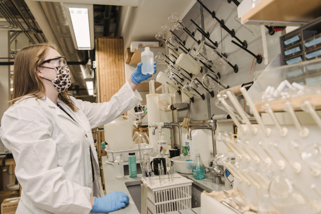 Woman in lab coat and mask looking at liquid in bottle