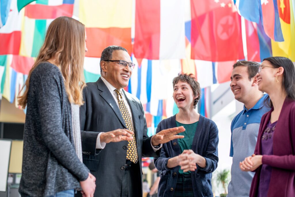 Older man in suit speaks with four smiling students, beneath international flags.