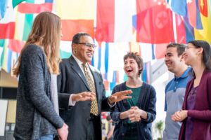 Older man in suit speaks with four smiling students, beneath international flags.