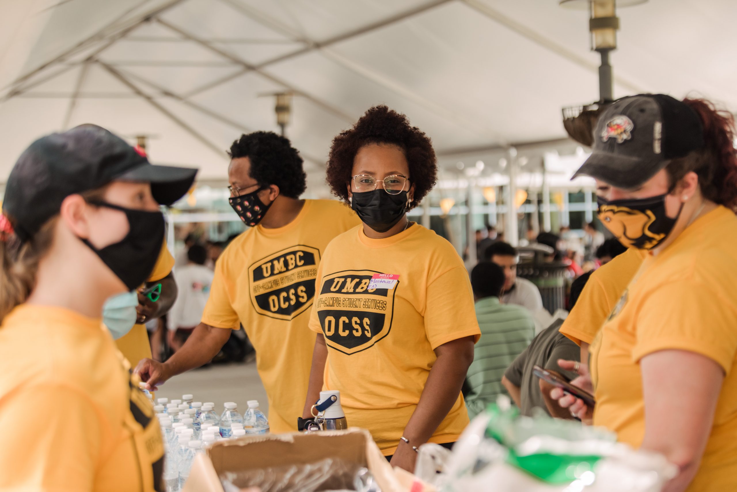 People wearing matching gold shirts are standing at a table. All the people are wearing black masks over their mouth and nose. One person in the center of the photo is looking at the camera.