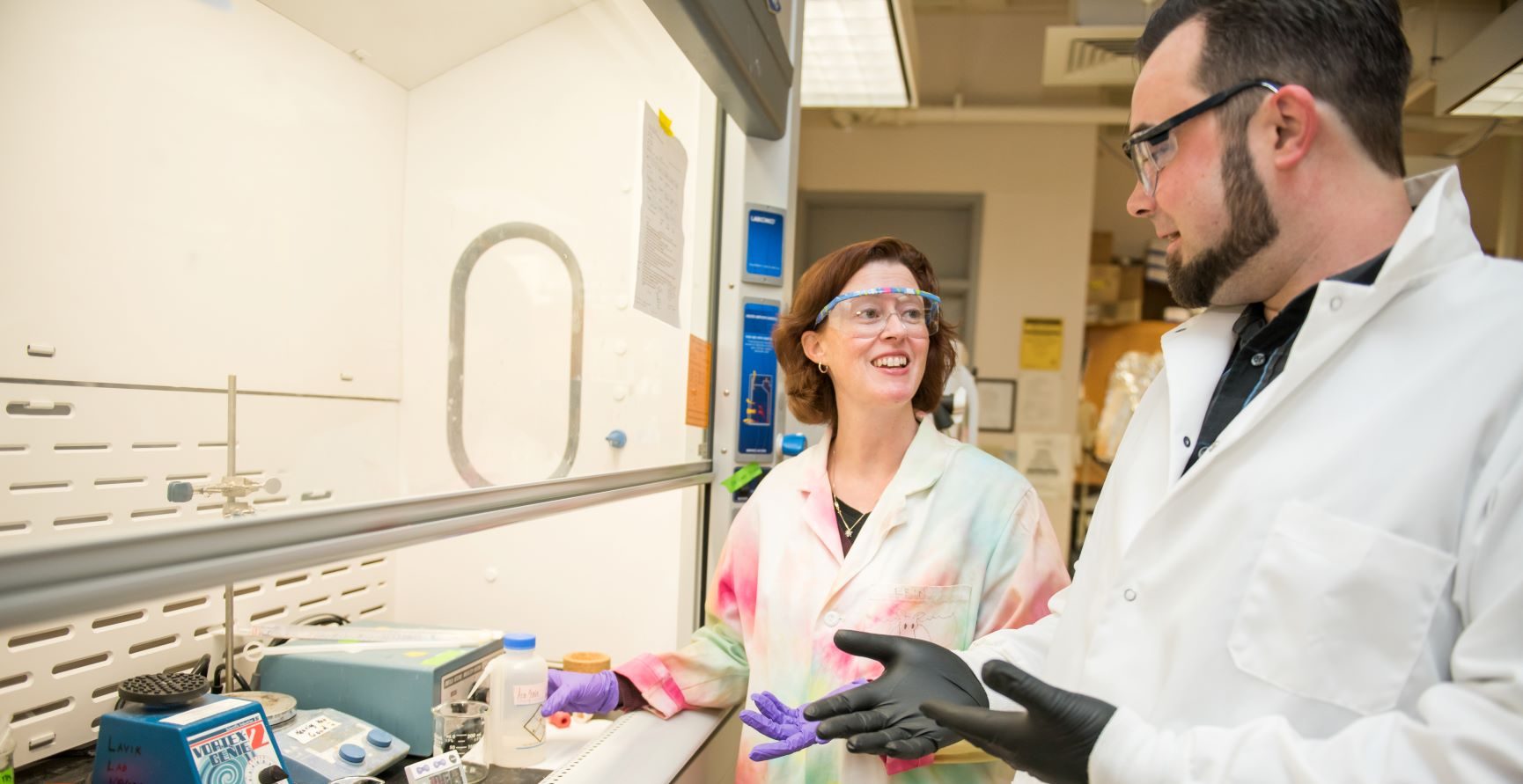 Woman with auburn hair stands with a man with dark bears in a lab. She wears a tie dyed lab coat and he wears a white lab coat.