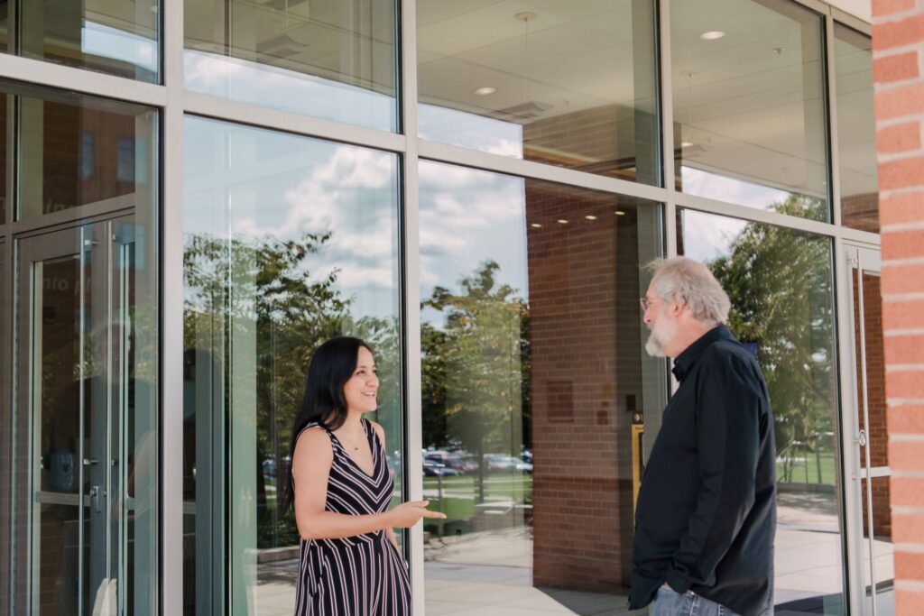 A woman and a man stand outside a glass and brick building facing each other.