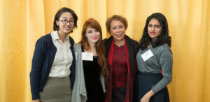 Four women stand together in front of a yellowbackground