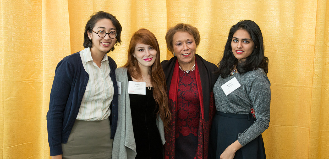 Four women stand together in front of a yellowbackground