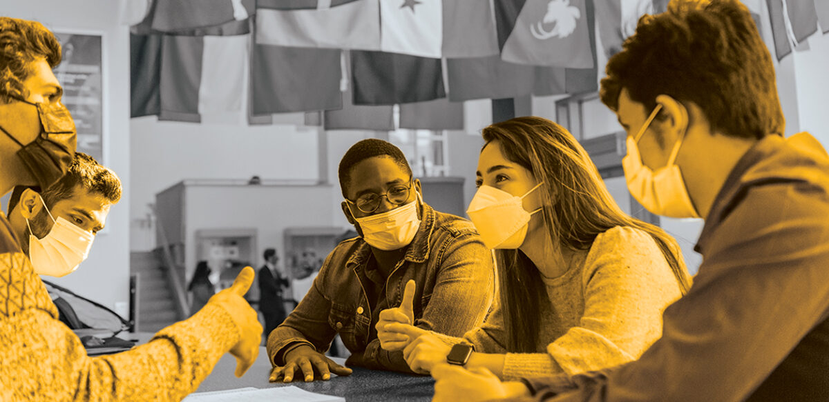 In gold overlay, students sit around a table in the commons talking.