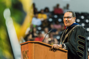 man in dark graduation robe stands behind a podium with positive expression