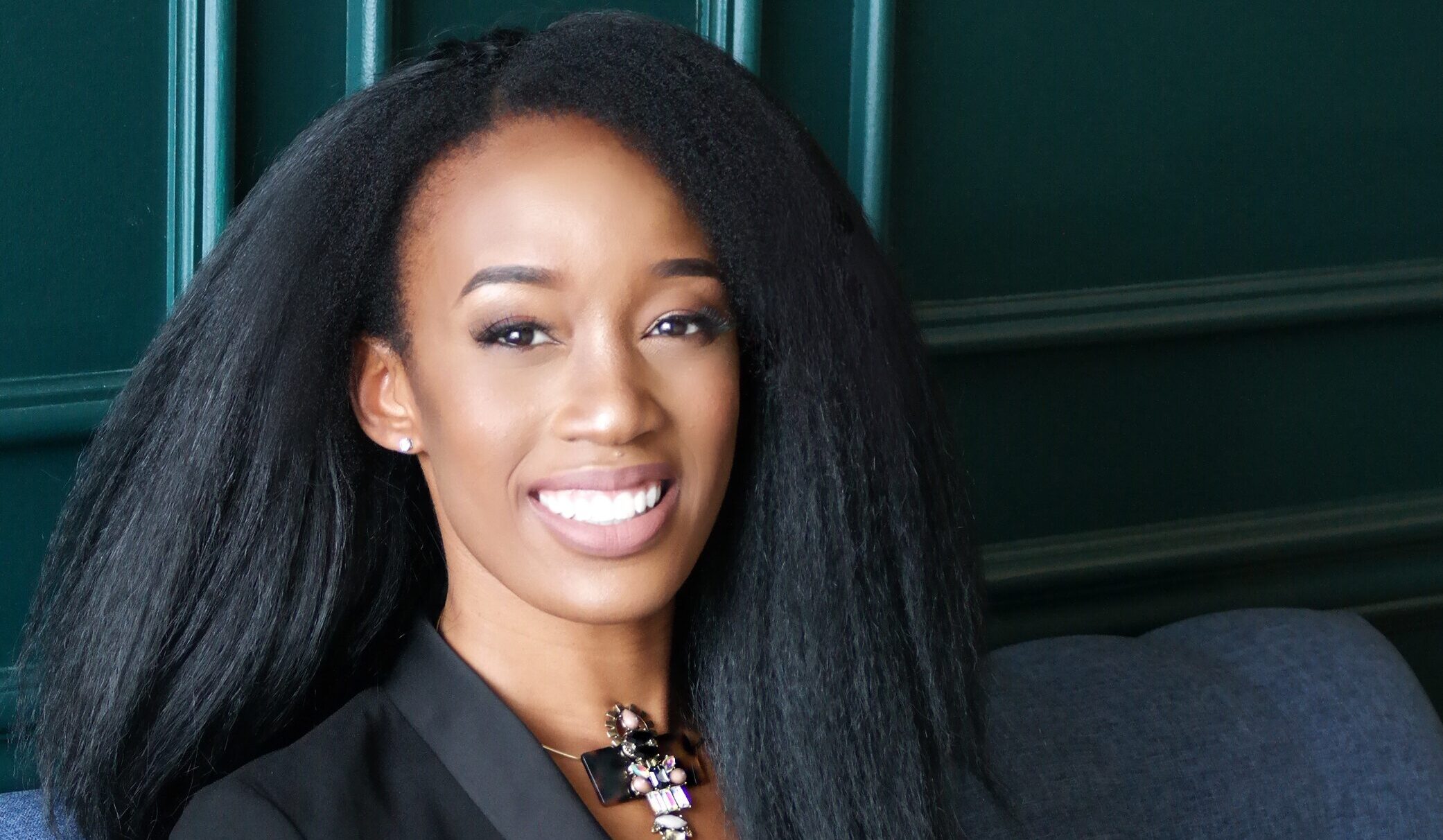 An adult with long, thick, black hair and wearing a black suit sits on a blue couch next to a small white pillow with the words life written on them in cursive Nkiru Nnawulezi