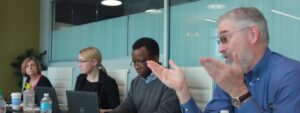 A group of five people in a room with glass walls, sitting at a table with laptops in front of them.