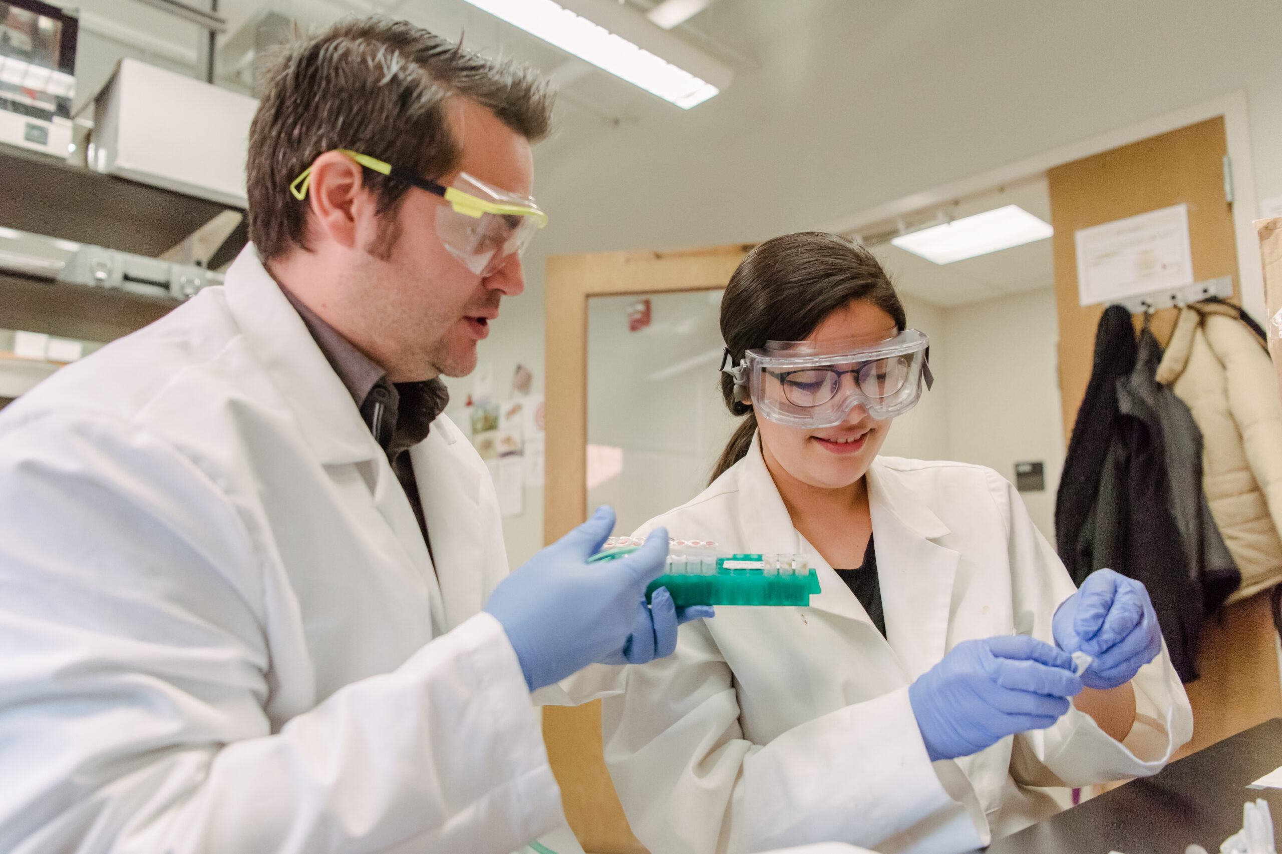 two people in lab coats and gloves examining small vials in a brightly lit lab