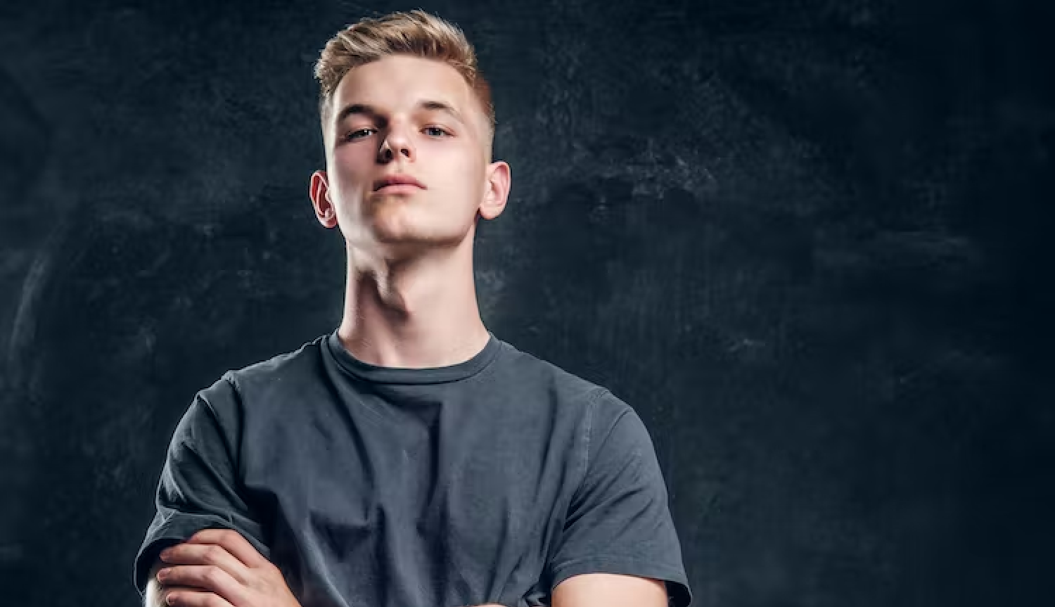 A person with cropped blond hair, wearing a grey t-shirt, stands with their arms crossed over their stomach, looking seriously. Democracy.