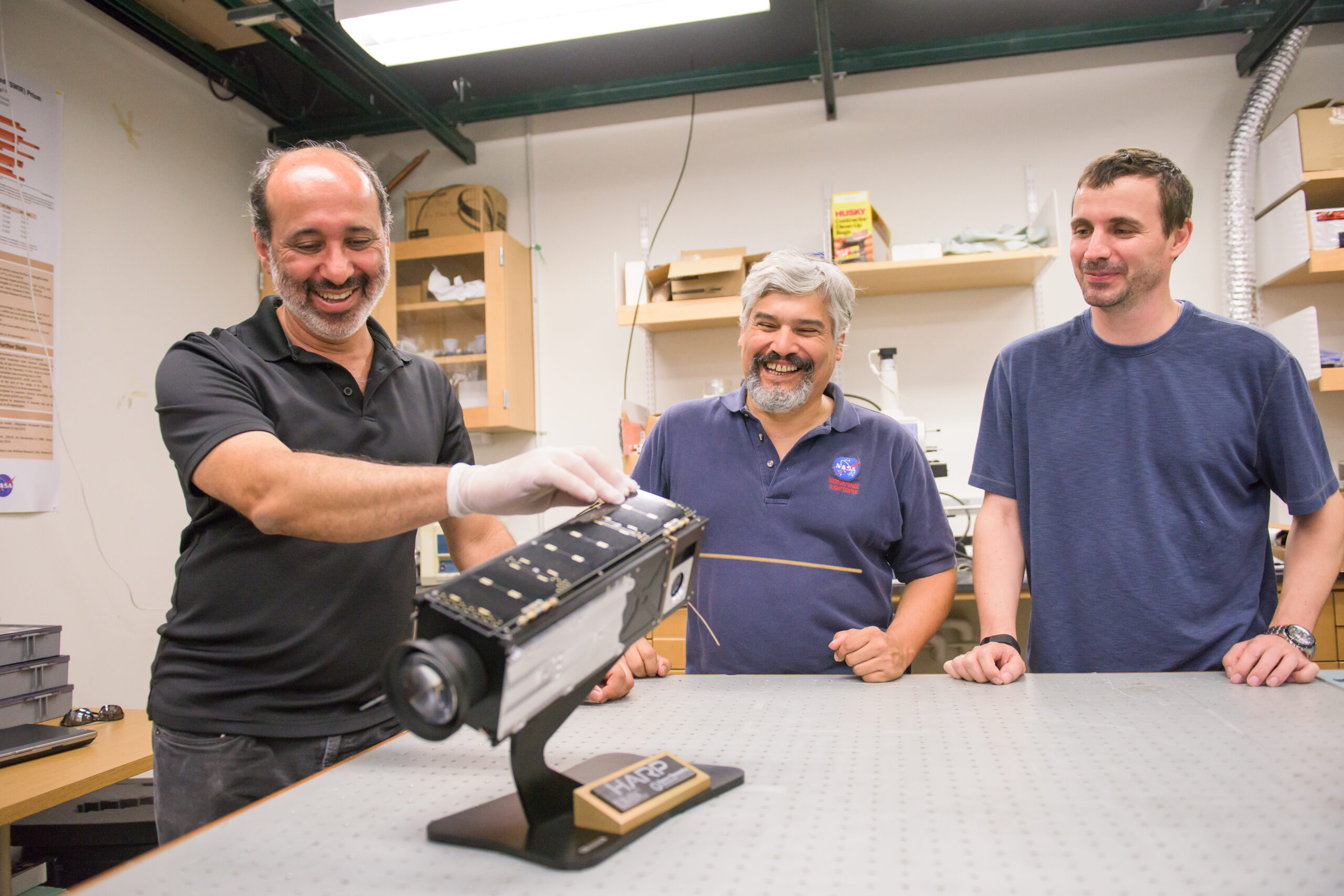 Three researchers stand around a table looking at the HARP cubesat on it (a mostly black rectangular solid on a simple stand)