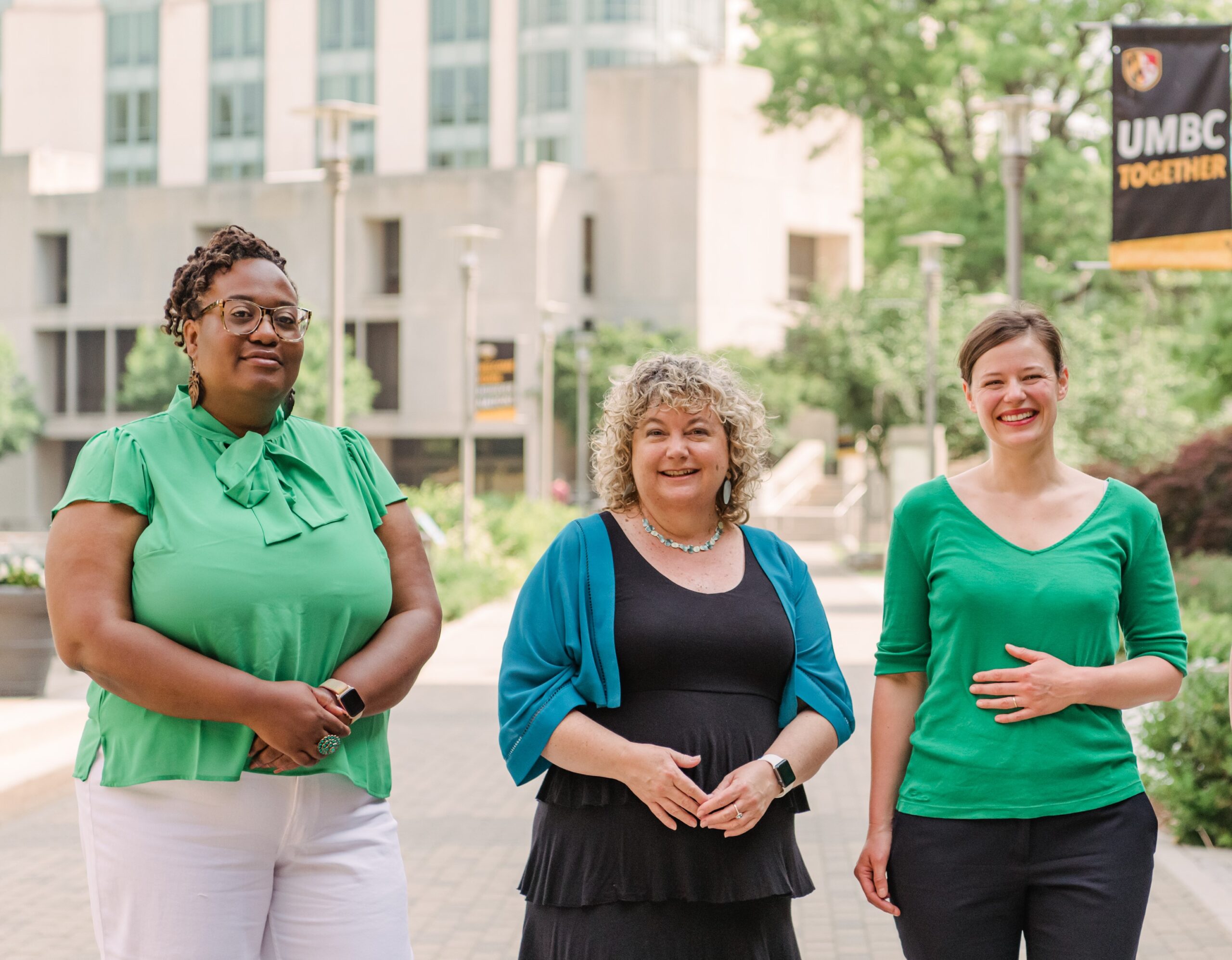 Three women stand side by side on a cement path with a white brick building behind them.