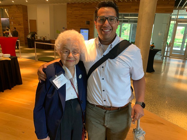 A young man with glasses stands with his arm around an older woman