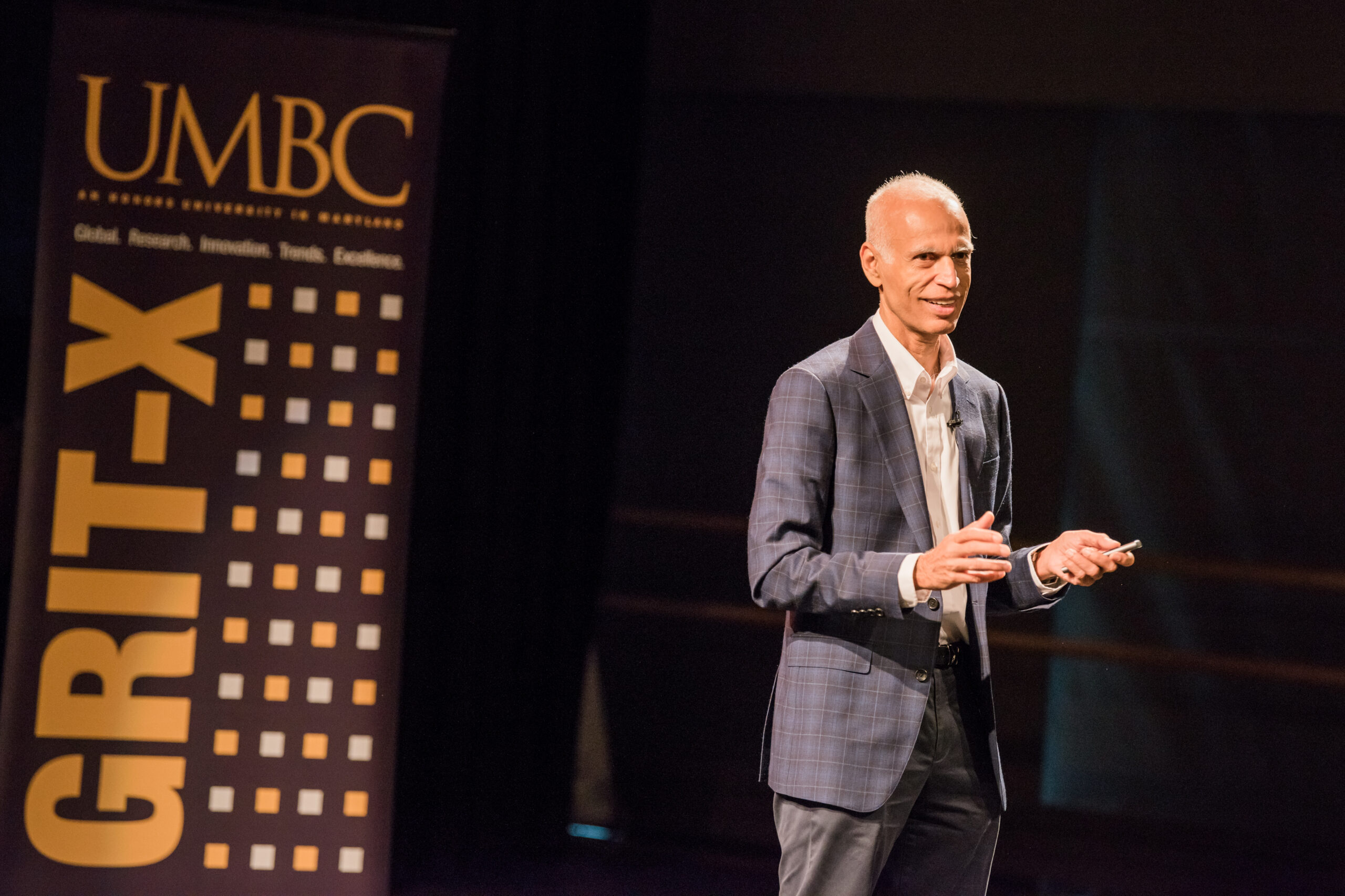 Man in a suit stands onstage in a dark theatre, with a vertical banner that says "UMBC - GRIT-X" behind him.