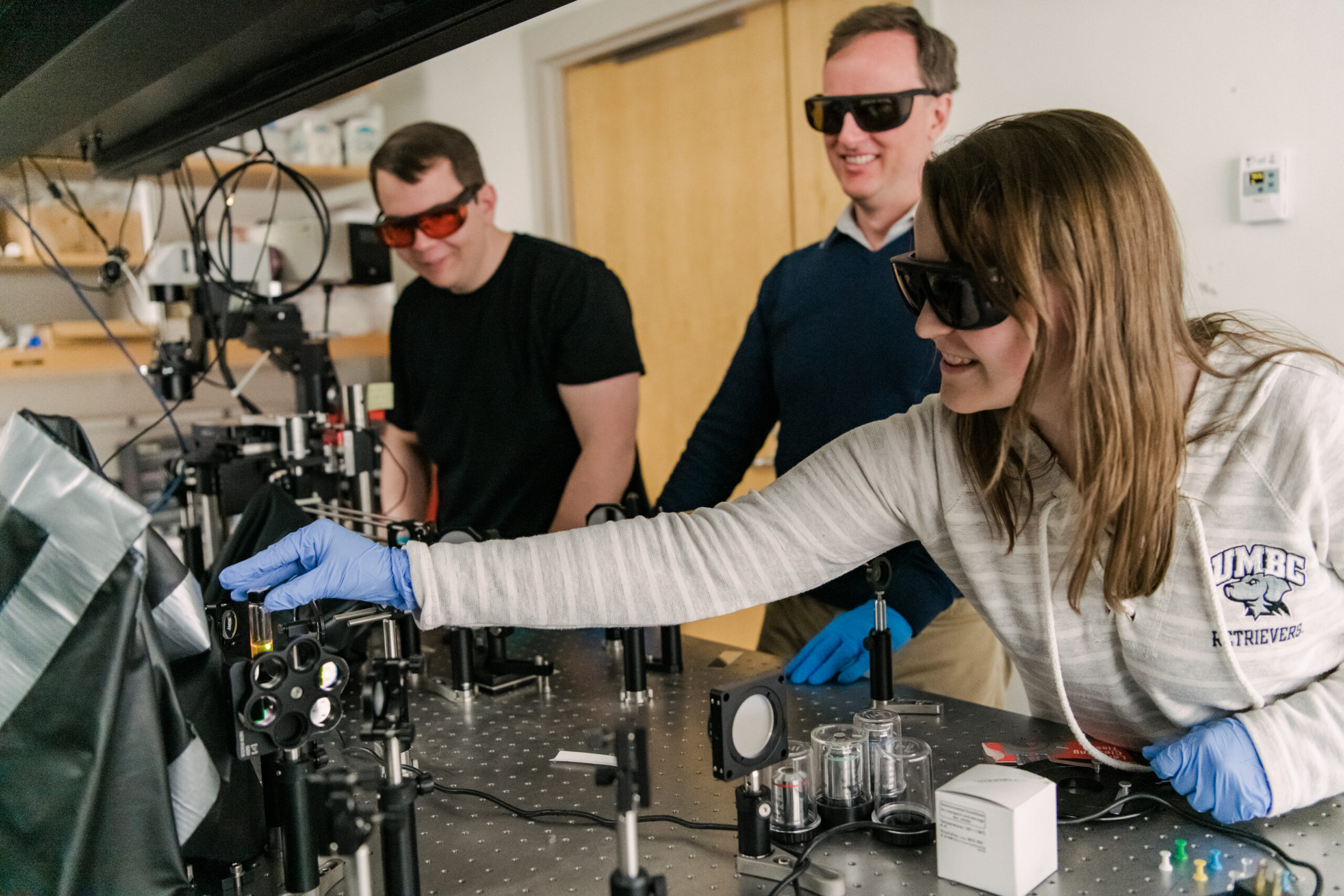 Three people work with machinery in a lab. They wear protective glasses and gloves. One wears a sweater reading "UMBC Retrievers."