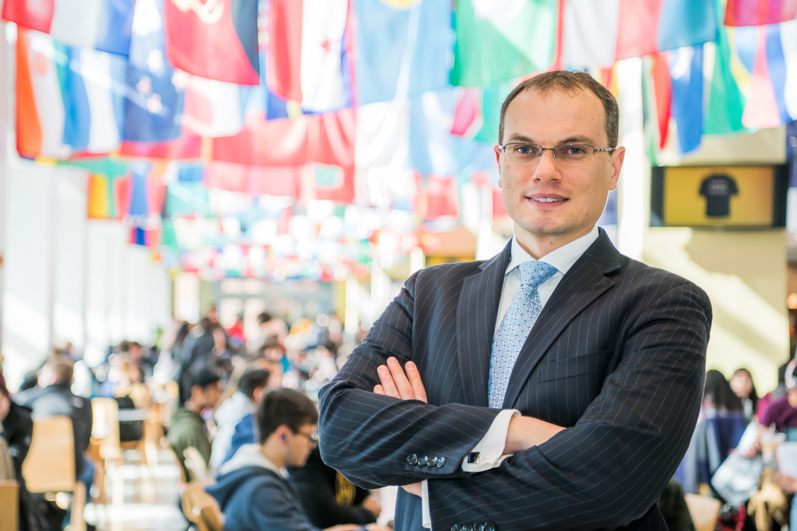 Man standing with his arms crossed in front of a backdrop of flags from around the world and students sitting.