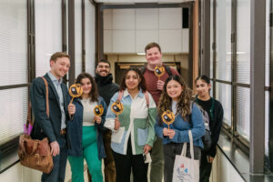 A group of students and a professor stand stand close together holding small gold and black signs, civic engagement