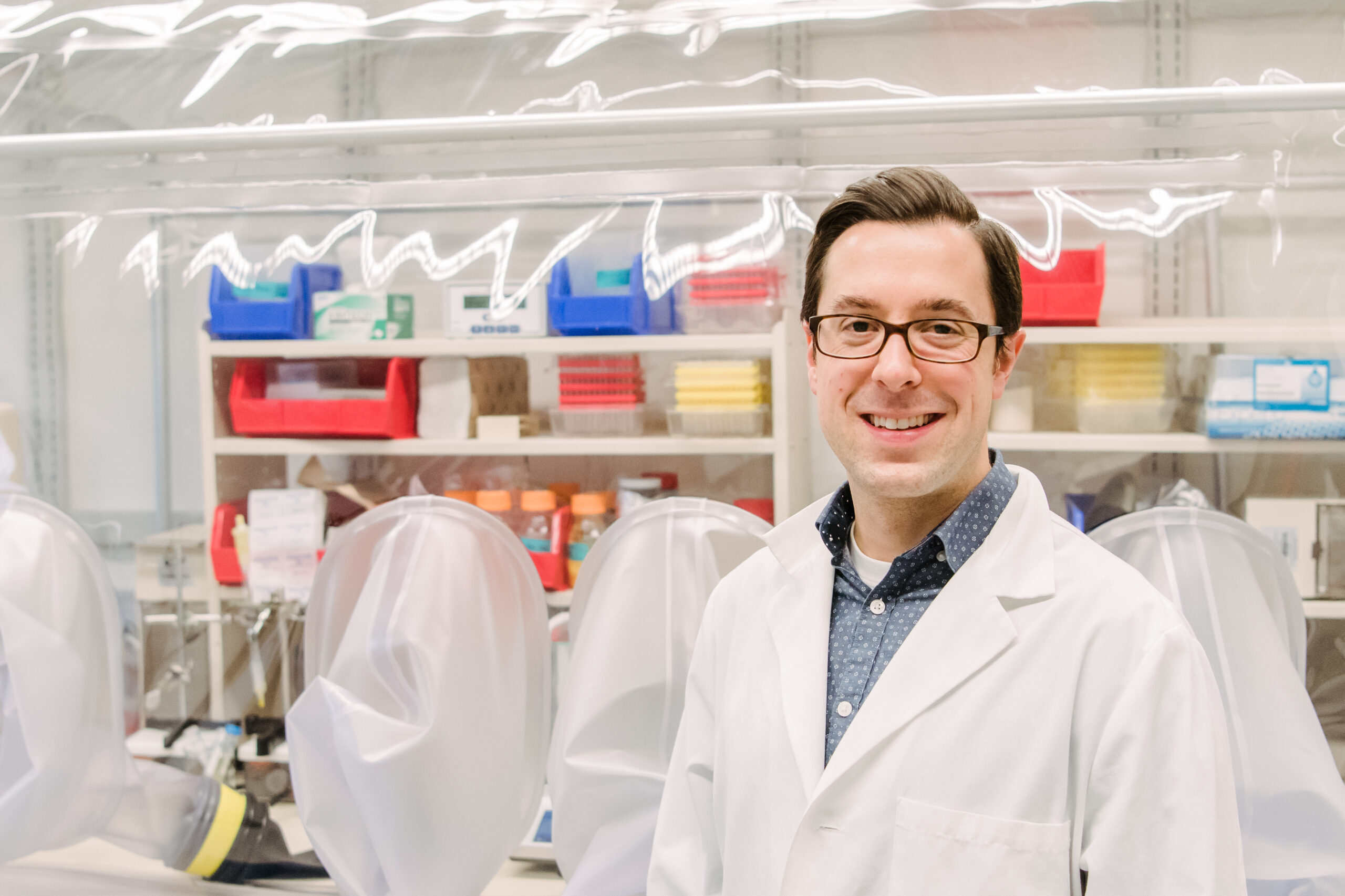 Man in white lab coat standing in front of a chemical hood with lots of colorful containers.