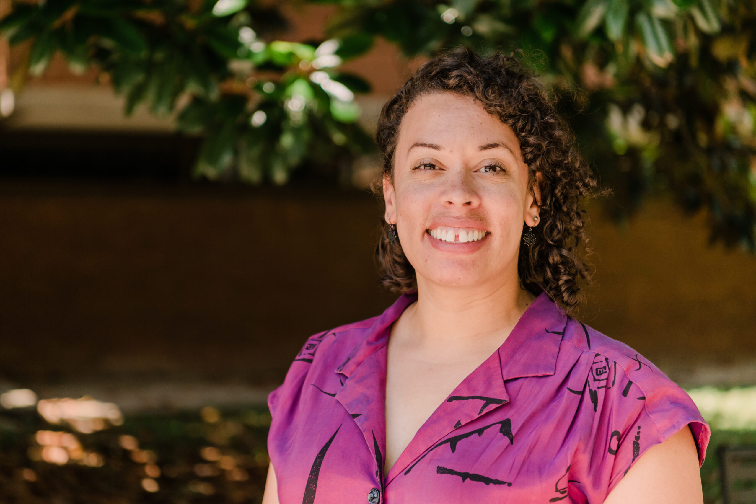 Portrait of a woman in a fuchsia top outside a brick building with some greenery.