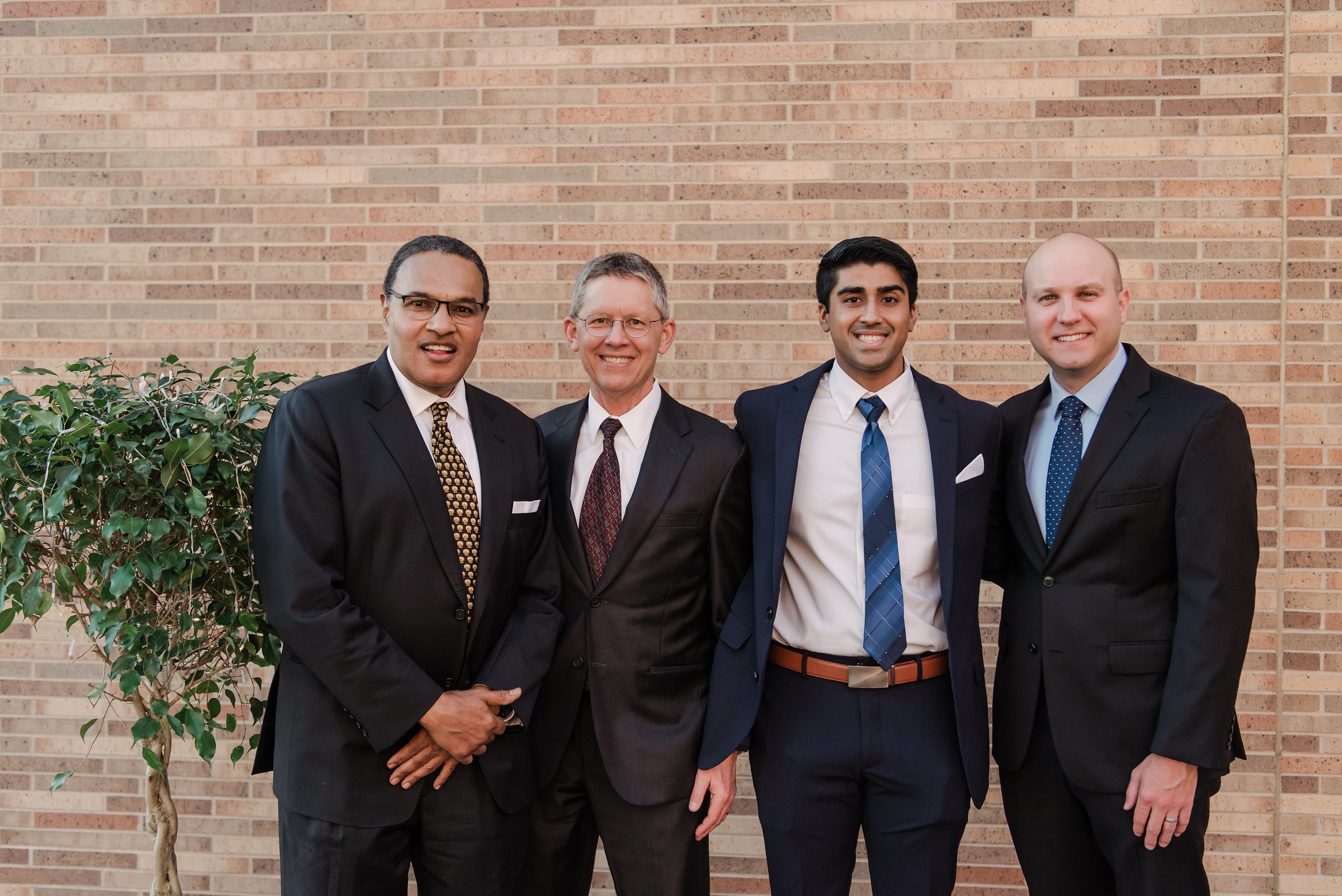 Four men wearing suits and ties pose together outside