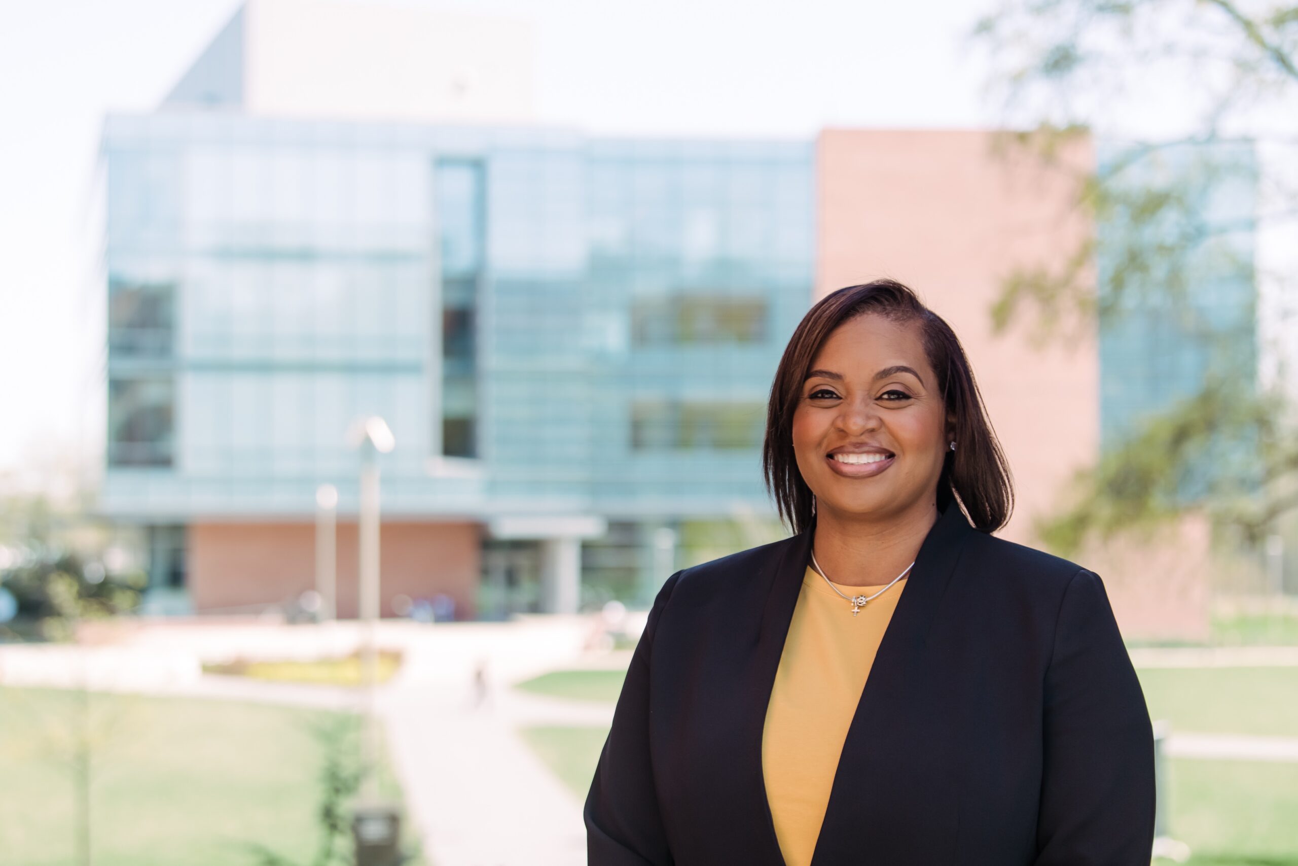 Tanyka M. Barber smiles for a professional headshot, wearing a yellow shirt and black blazer, outdoors, in front of a larger glass and brick building