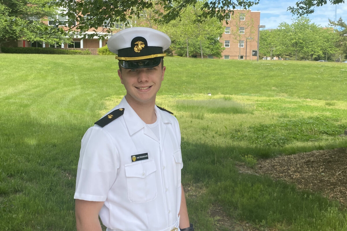 Man in white uniform stands in front of lawn, with buildings in background.