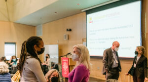 In the foreground and background two groups of two people stand in a crowded room talking.