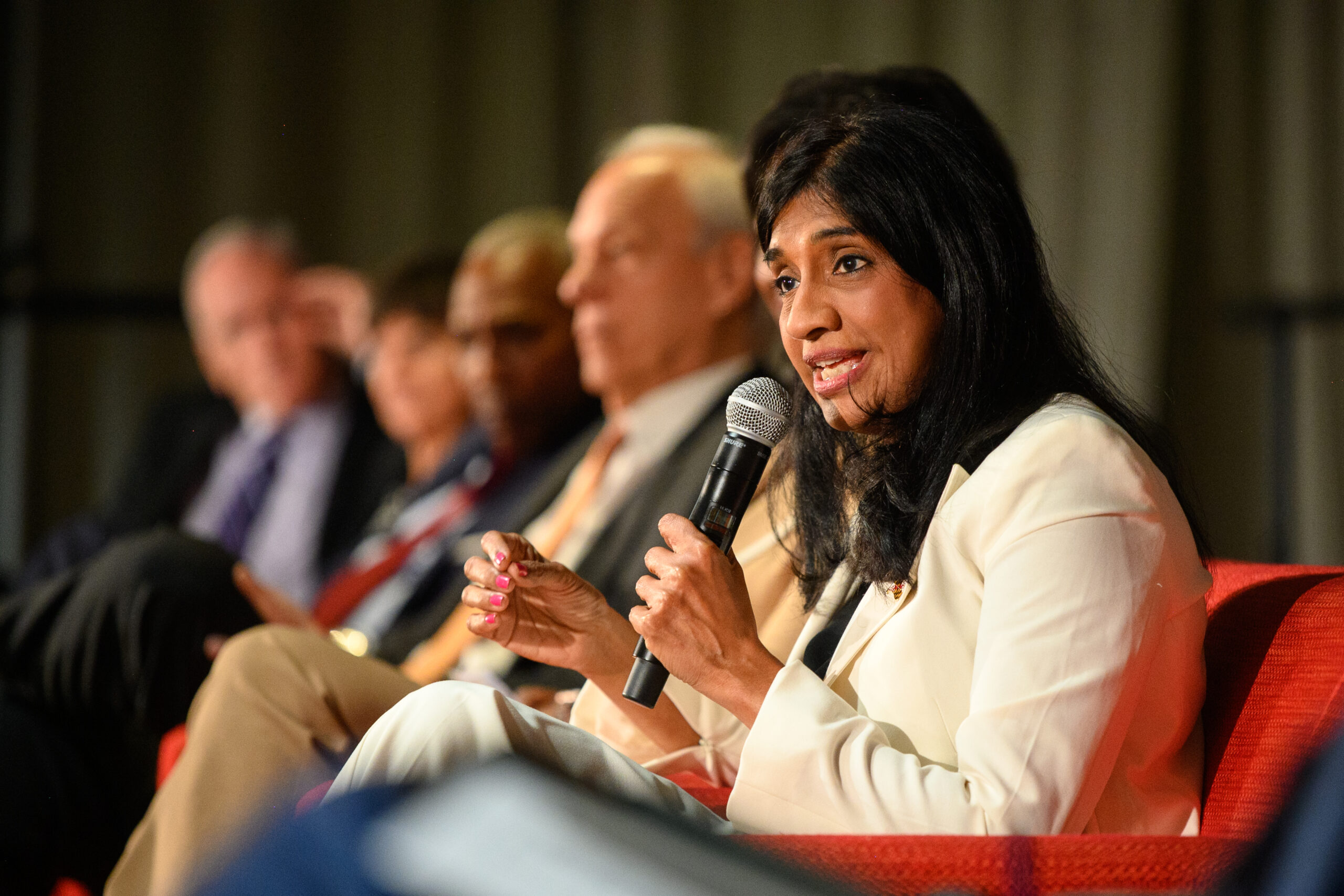 Woman in a cream-colored suit speaks into a microphone while seated in a red chair. Additional people in professional clothing are seated in a row next to her.