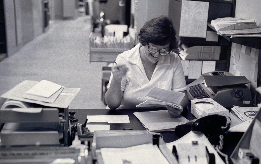 in a black and white photo, a woman sits at a desk, pouring over materials