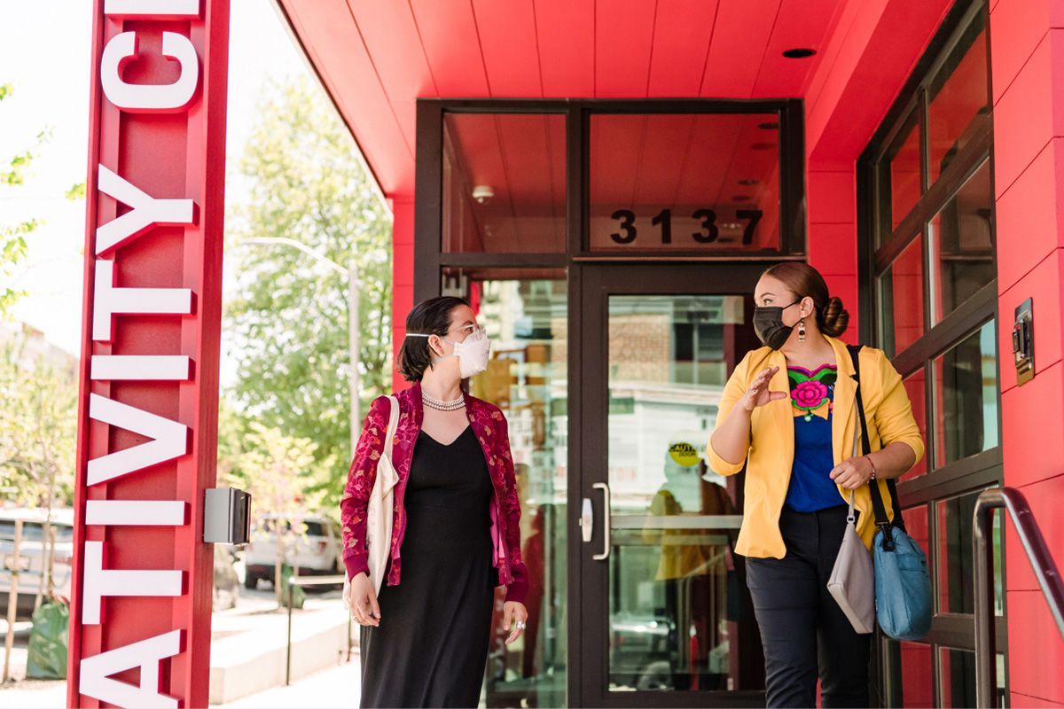 Two women wearing a COVID-19 and red and yellow blazers, walk down a set of steps in front of a red building.