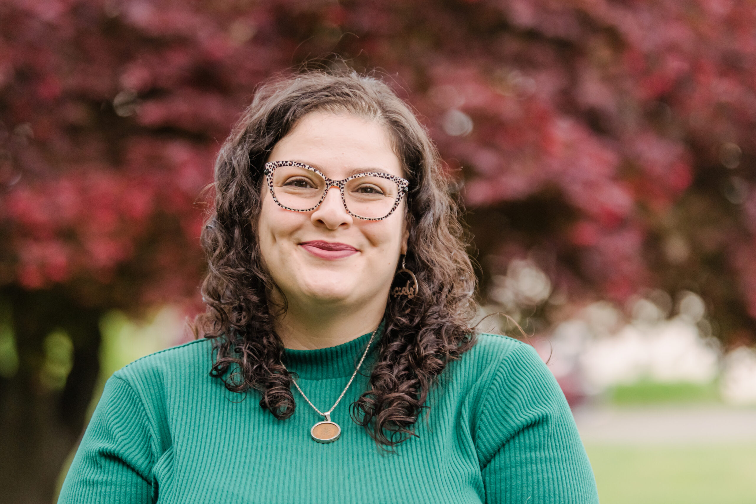 A woman with long curly brown hair wearing a green sweater stands outside with a red maple tree in the background. Arab
