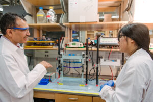 two scientists in lab coats converse at a lab bench with equipment on it