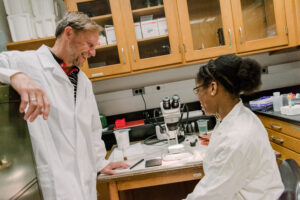 two people in lab coats, one sitting, one standing, having a conversation at a lab bench.