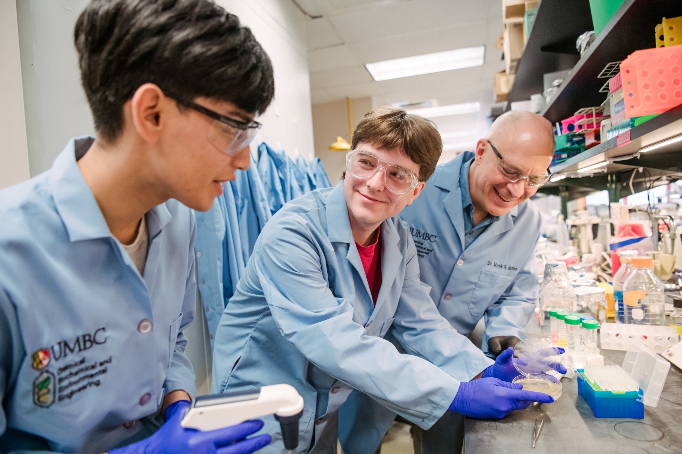 Three people in lab coats examine equipment at a lab bench.