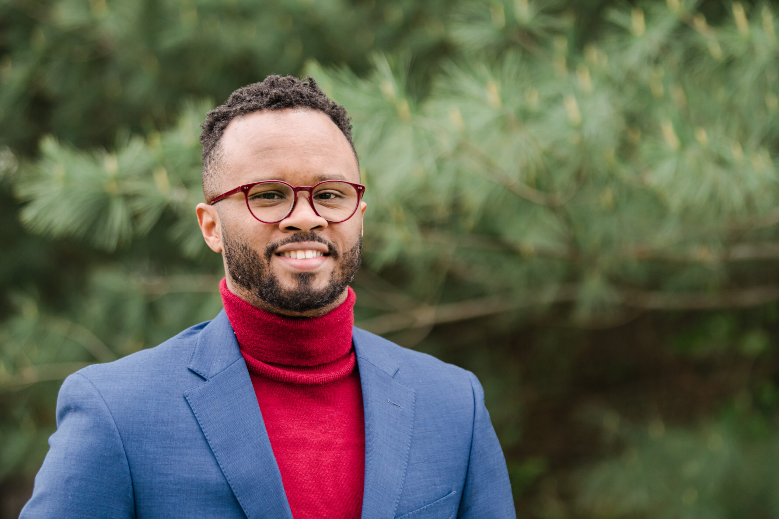 Ramon Goings wearing a blue blazer and red turtle-neck stands outside in front of large pine trees. Academic Minute