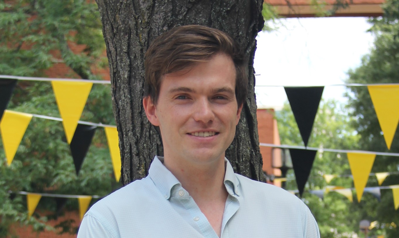 An adult wearing a light blue collar shirt stands outside with black and gold banners and trees behind them. Hilltop Institute