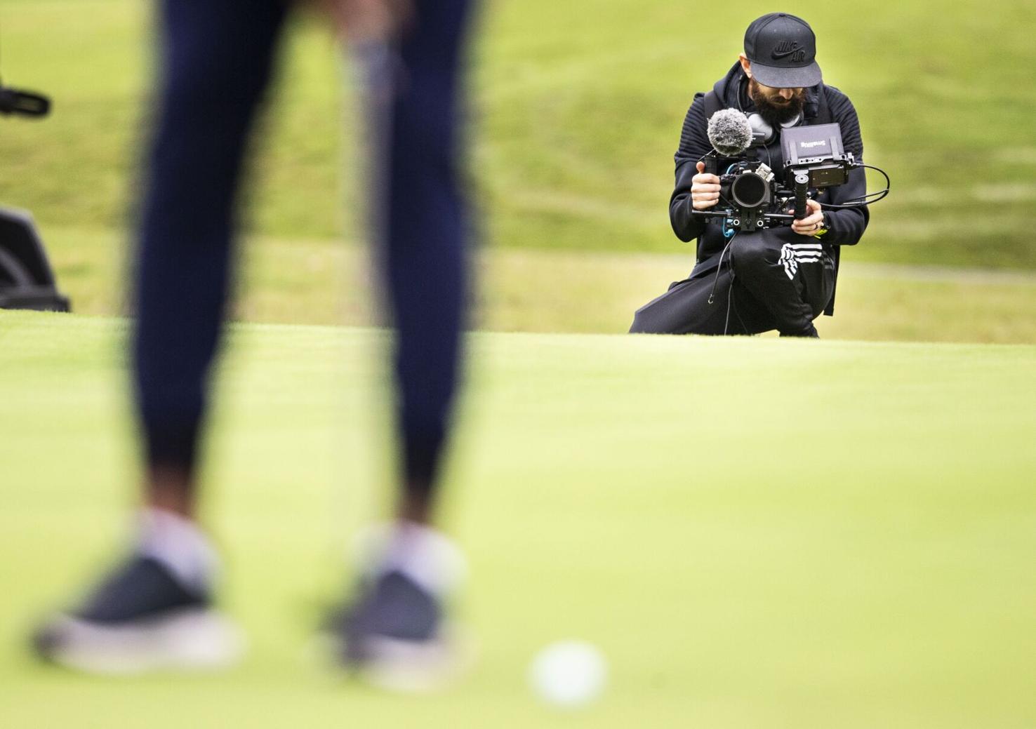 on a golf green, a man kneels with a camera, filming some one playing golf