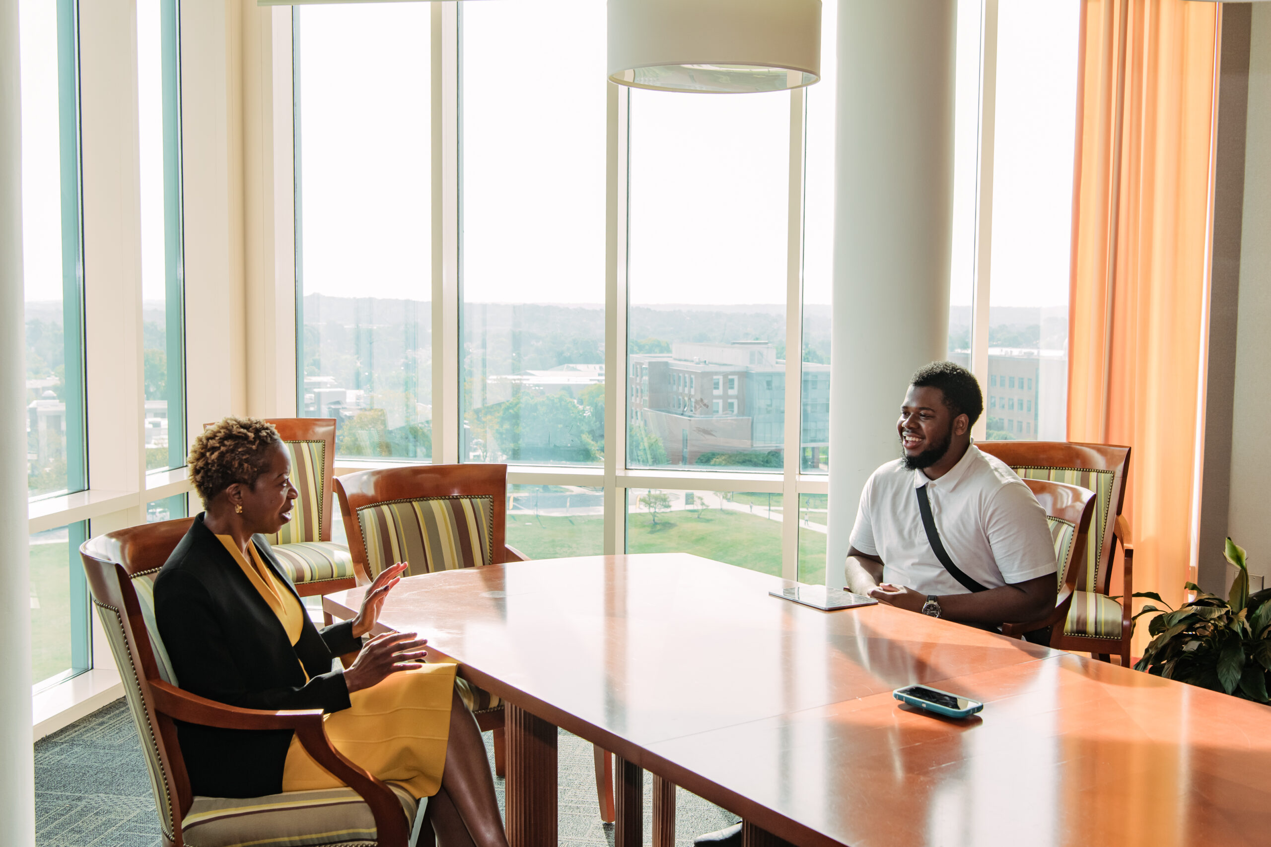 A woman and a college student sit talking to each other across a conference table with lots of windows behind them during office hours
