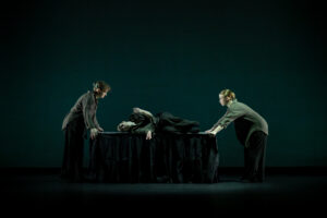 Two dancers stand on opposite sides of a table while a dancer lays on it between them