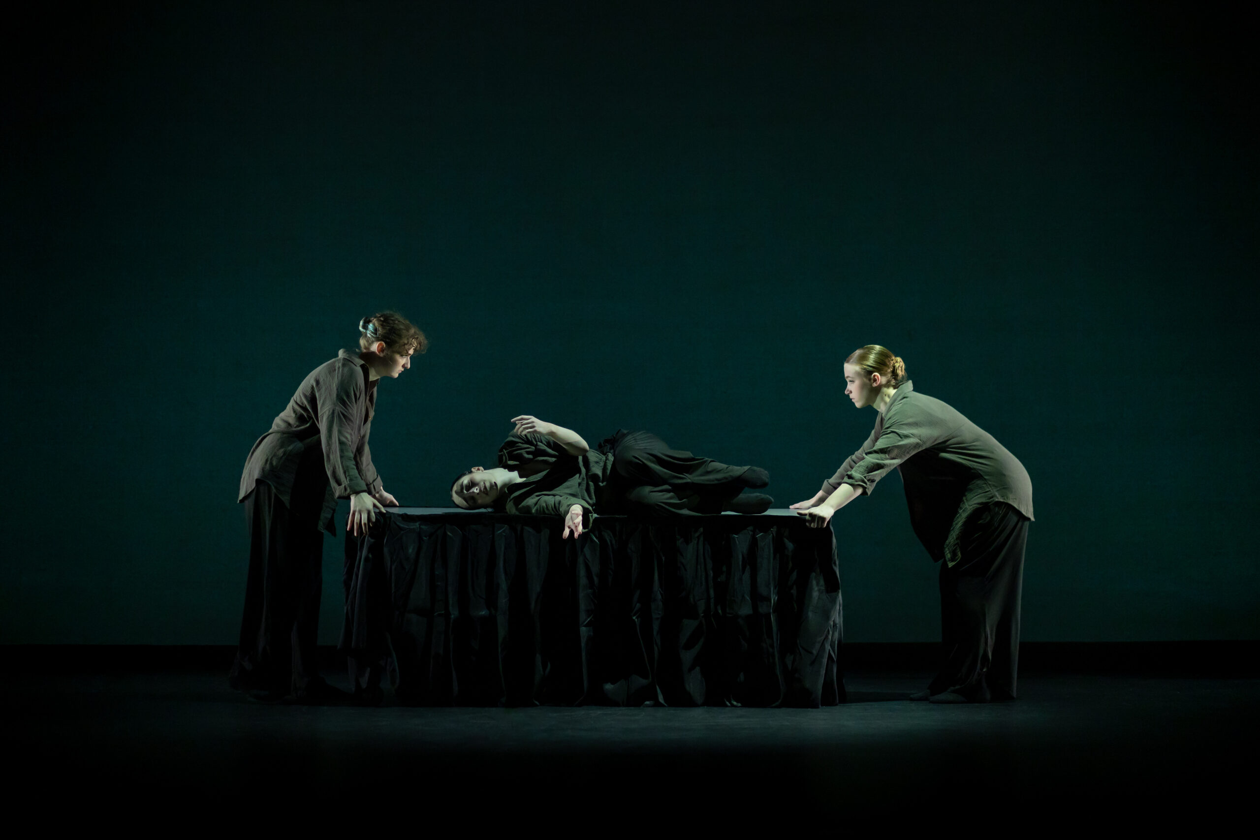 Two dancers stand on opposite sides of a table while a dancer lays on it between them