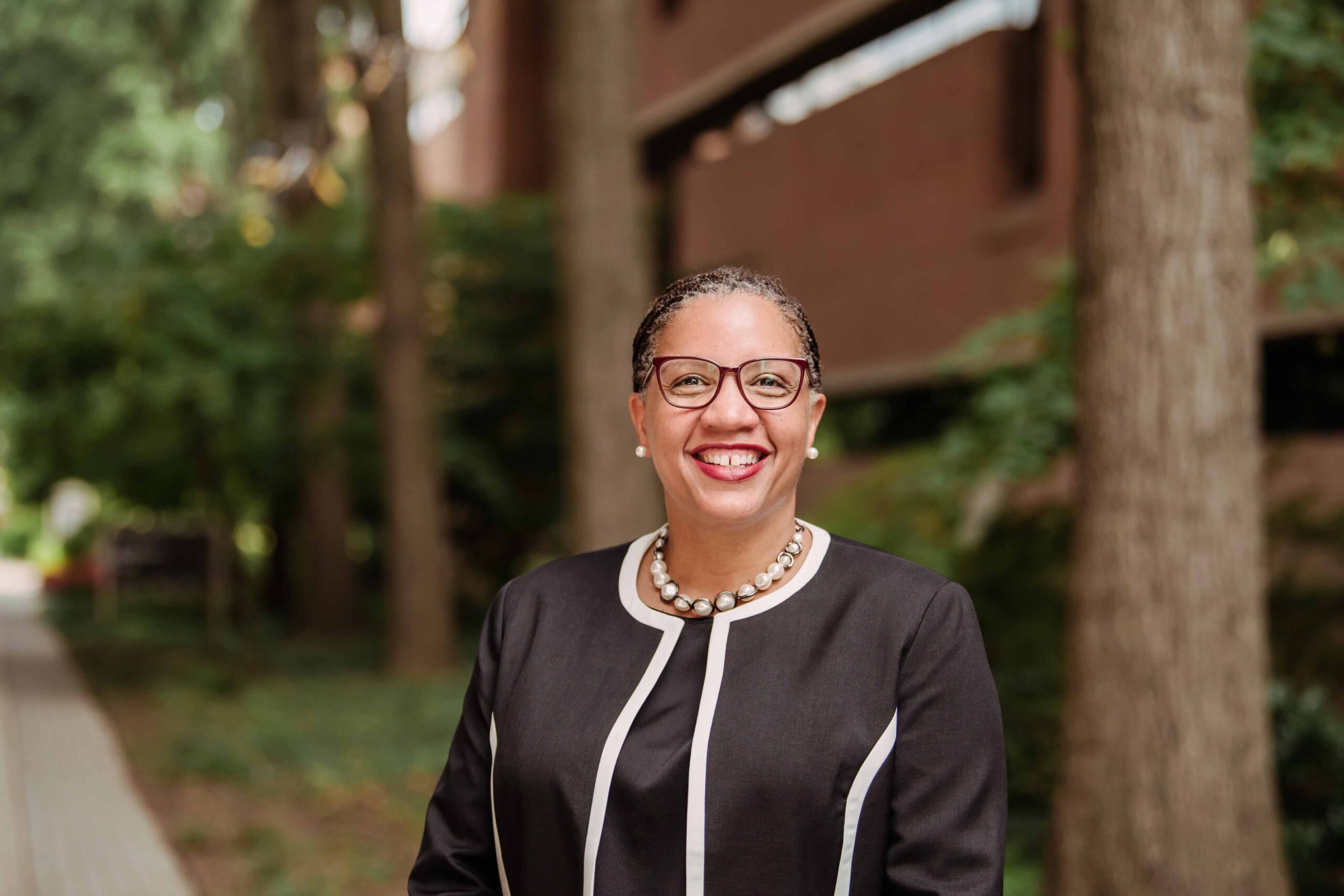 a woman in glasses stands in a business suit against a wooded background
