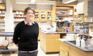 portrait of woman wearing a black sweater in a laboratory, backed by lab counters and shelving with chemicals and flasks