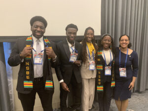 group photo of five people in professional attire and conference lanyards, two wearing black stoles with red, green, and yellow accents.