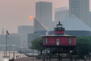 Smoky skies and an orange sun backdrop skyscrapers near a harbor.